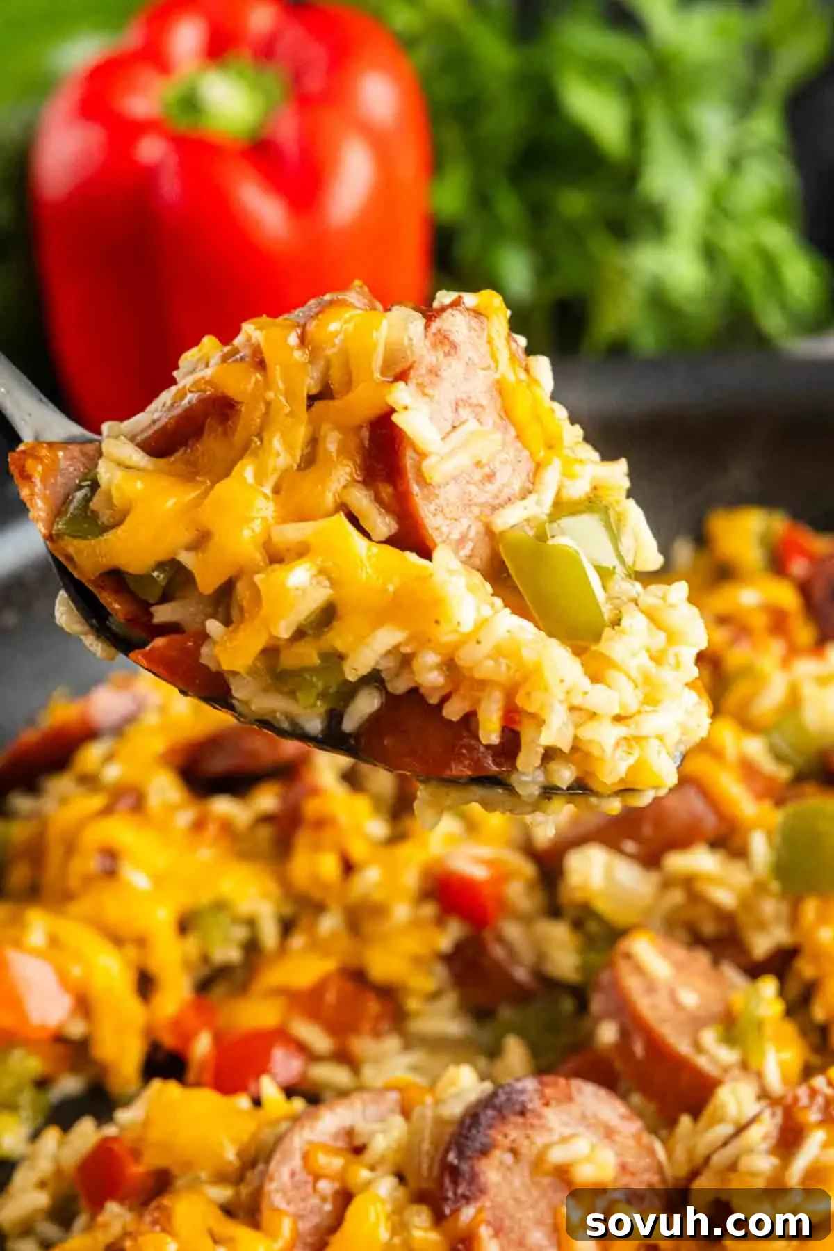 A spoonful of cheesy sausage and rice with savory sausage, bell peppers, and melted cheddar, with a red bell pepper and greens in the background.