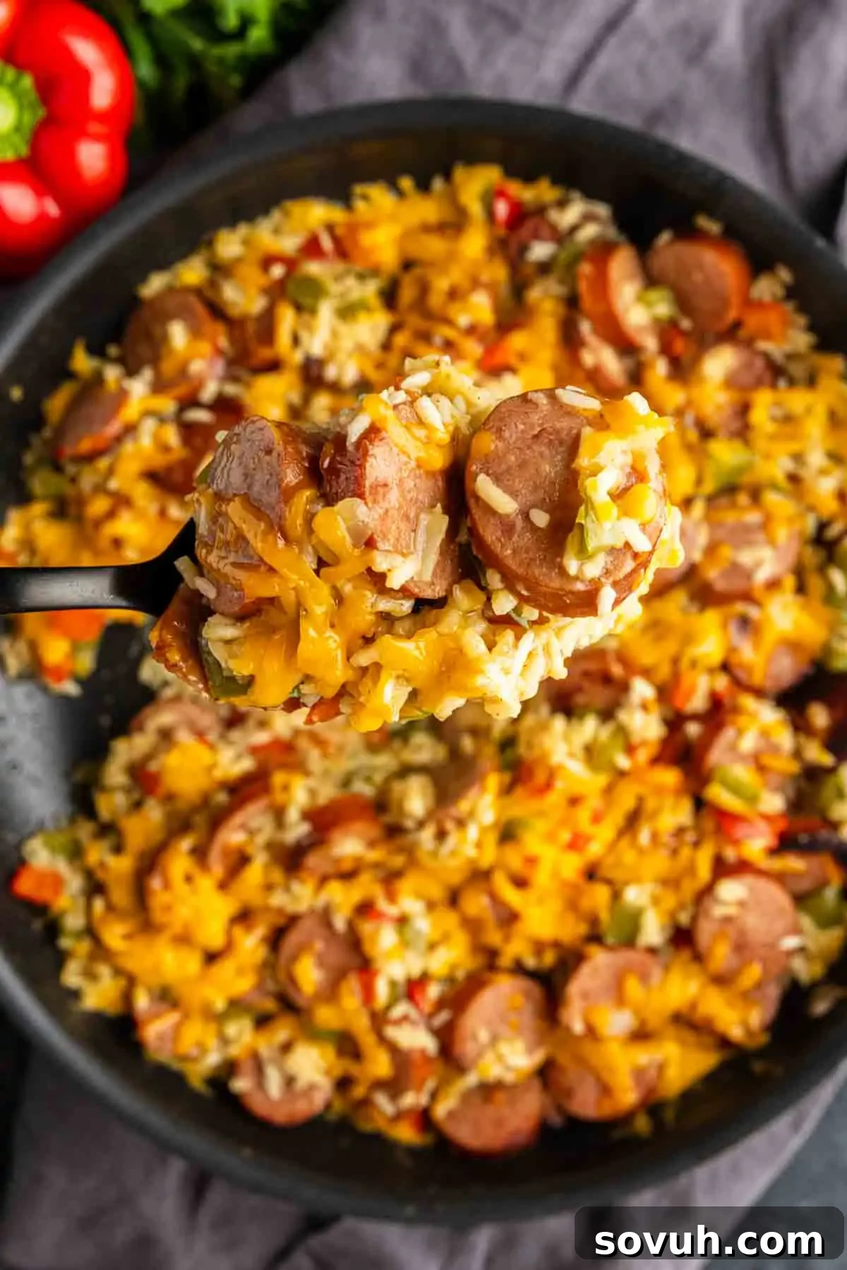 A close-up of a one pot cheesy sausage and rice skillet filled with sausage, rice, bell peppers, and melted cheese, with a spoon lifting a portion from the pan.