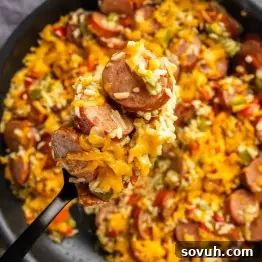 A close-up of a spoonful of sausage, rice, chopped vegetables, and melted cheddar cheese, with more of the same dish in the background.