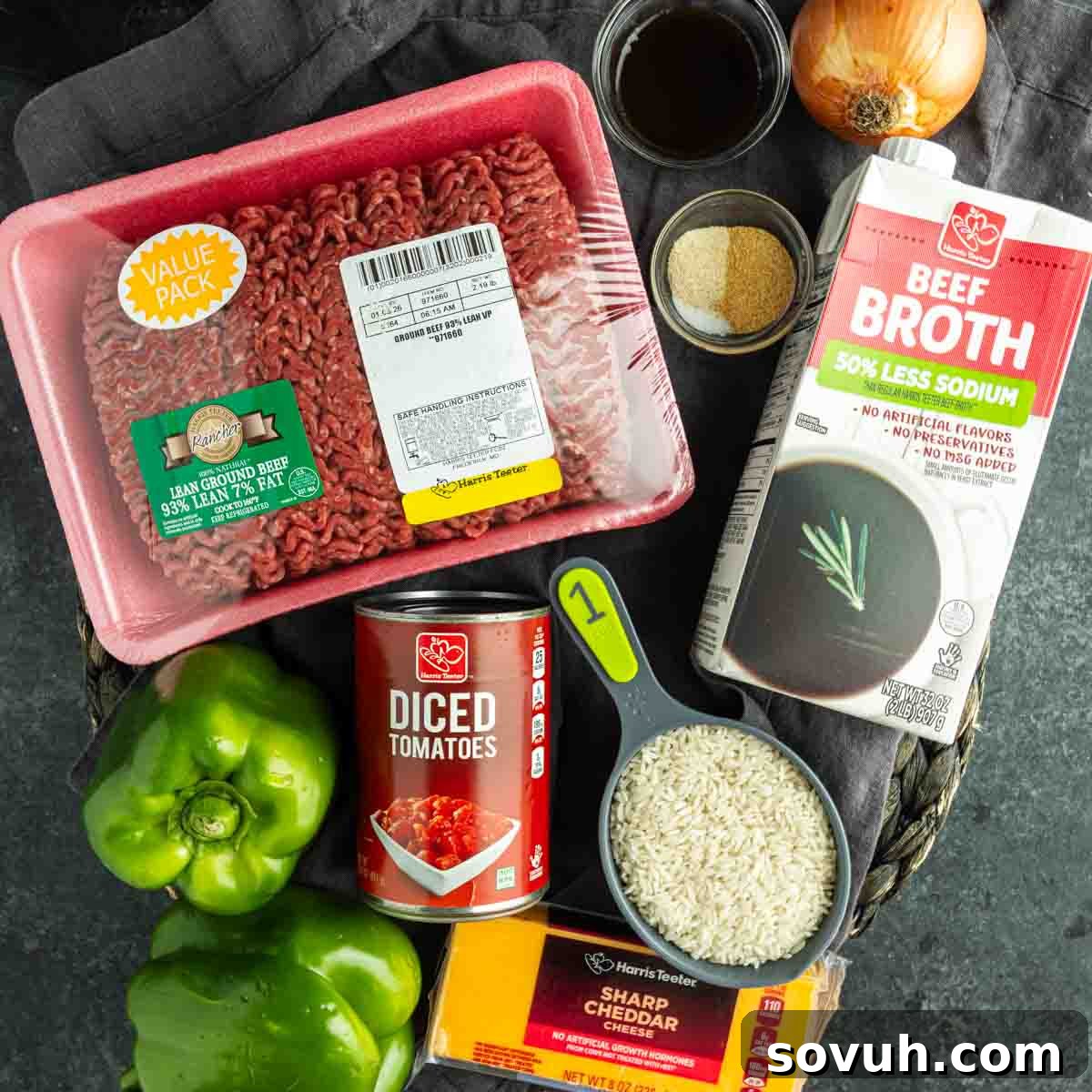 Top-down view of fresh ingredients for a ground beef and rice skillet—including beef broth, diced tomatoes, vibrant green bell peppers, shredded cheddar cheese, onion, and various seasonings—beautifully arranged on a dark cutting board, ready for an easy unstuffed pepper skillet meal.