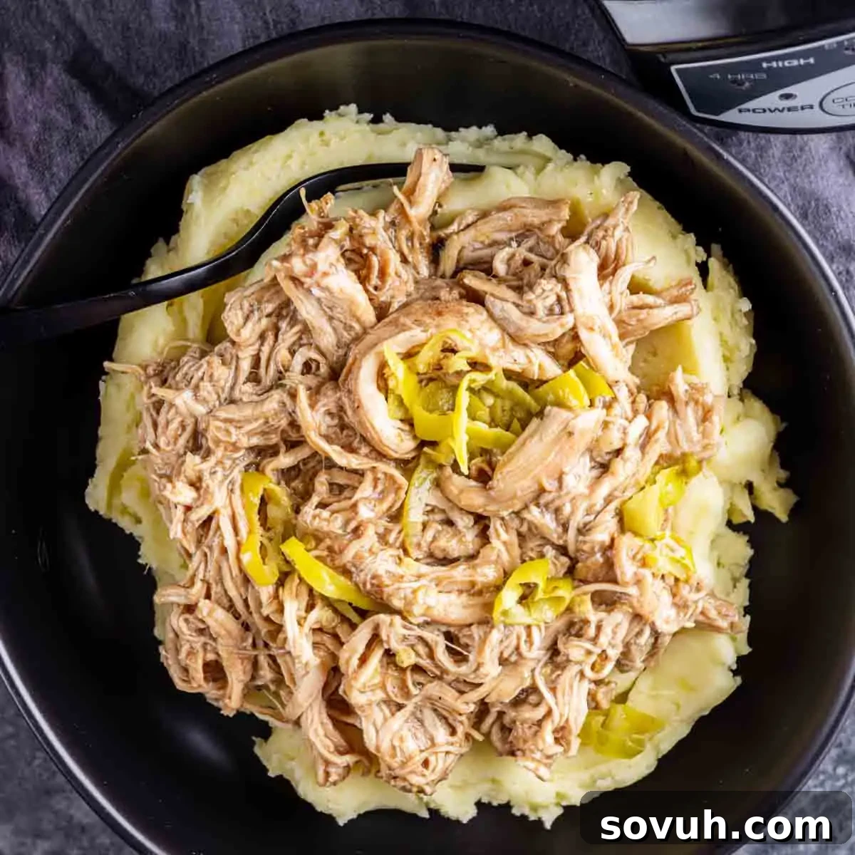 A black bowl filled with mashed potatoes topped with Slow Cooker Mississippi Chicken and sliced yellow peppers. A black spoon rests in the bowl.
