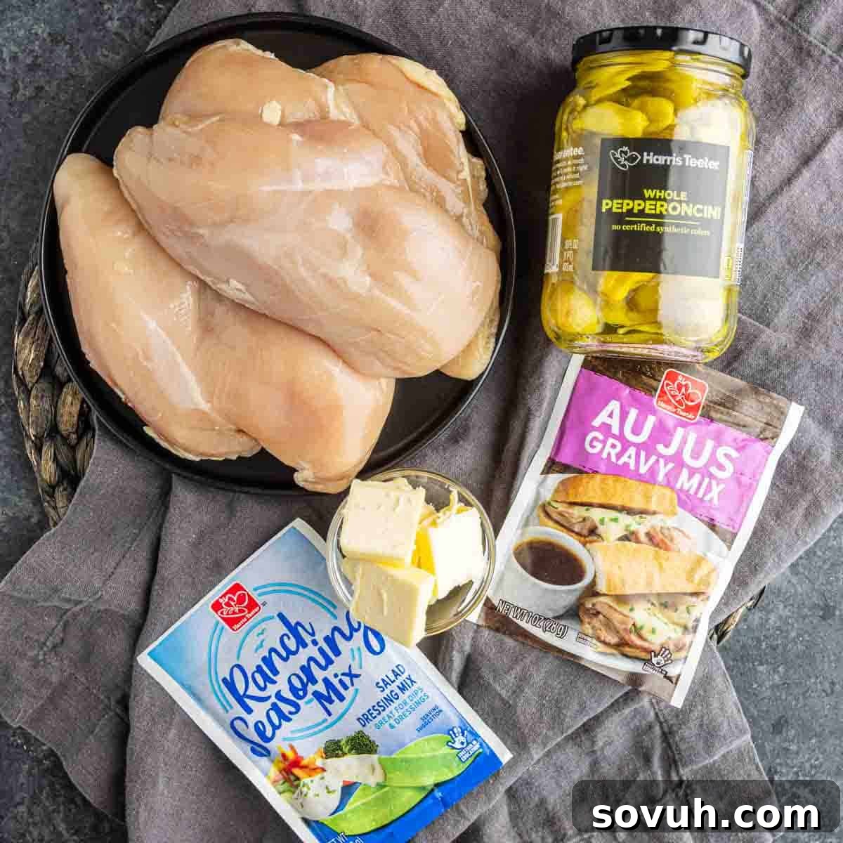 Raw chicken breasts, whole pepperoncini, ranch seasoning, au jus gravy mix packets, and butter on a plate—all you need for an easy dump and go crockpot meal like Slow Cooker Mississippi Chicken.