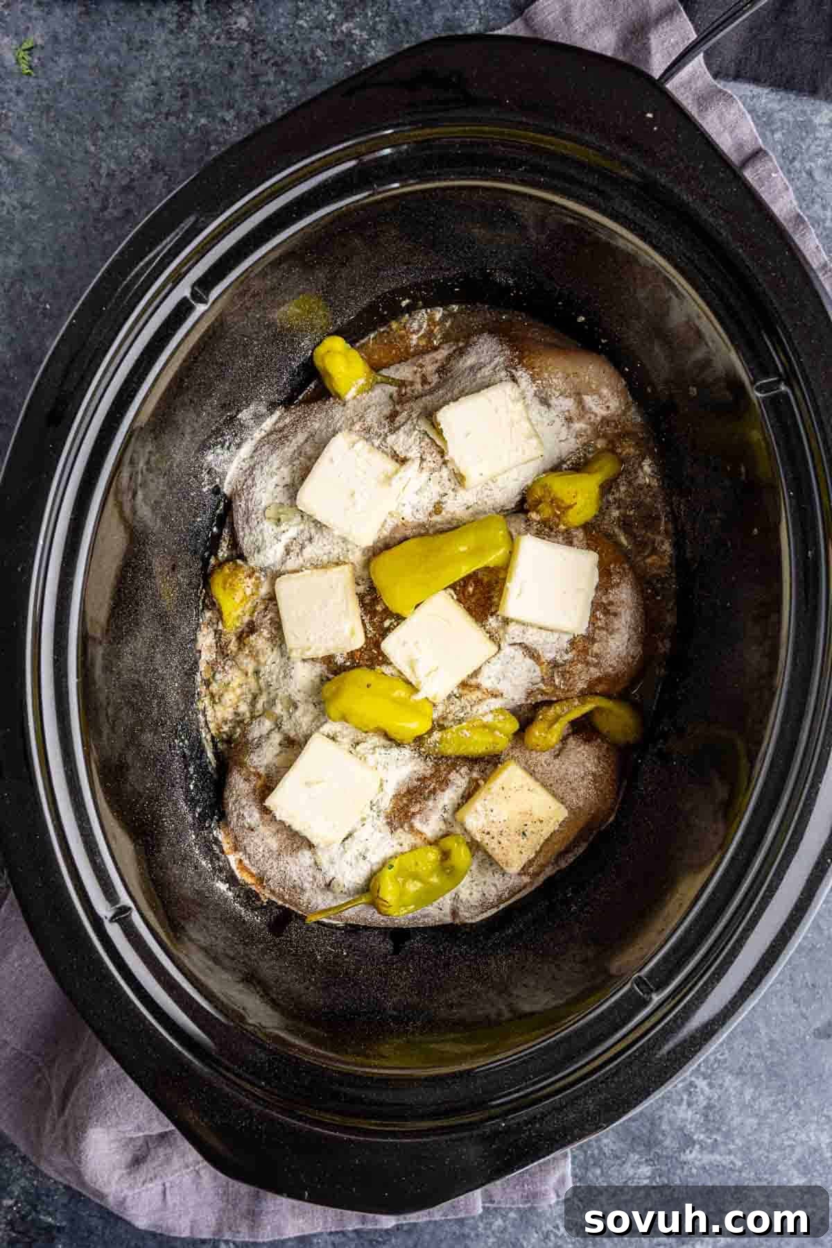 Three raw beef roasts in a slow cooker topped with sliced butter, pepperoncini peppers, and seasoning—an easy dump and go crockpot meal ready to be cooked.