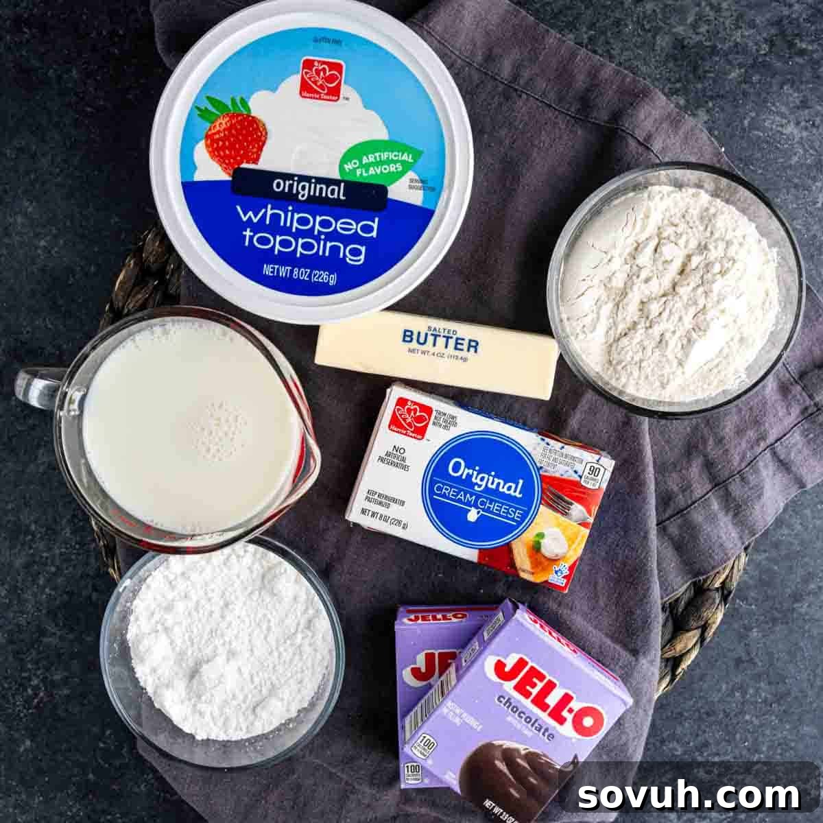 Decadent Chocolate Dream 3 Ingredients including whipped topping, flour, stick of butter, milk, cream cheese, powdered sugar, and chocolate pudding mix arranged on a dark surface—perfect for making a rich layered chocolate pudding dessert like Chocolate Delight.