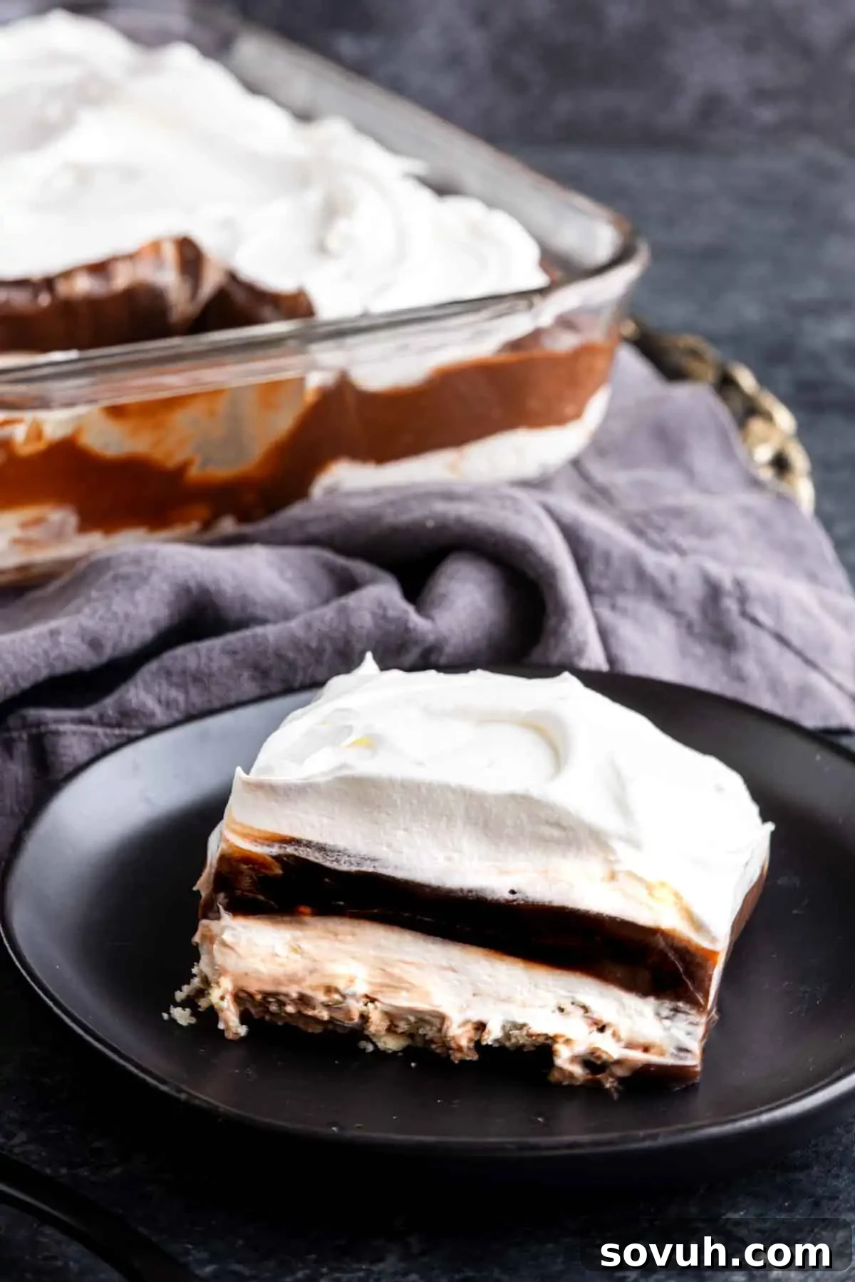Decadent Chocolate Dream 6 A slice of luscious chocolate lasagna dessert with whipped cream, rich chocolate, and cream filling sits on a black plate, while the rest of the layered treat awaits in a glass dish in the background.