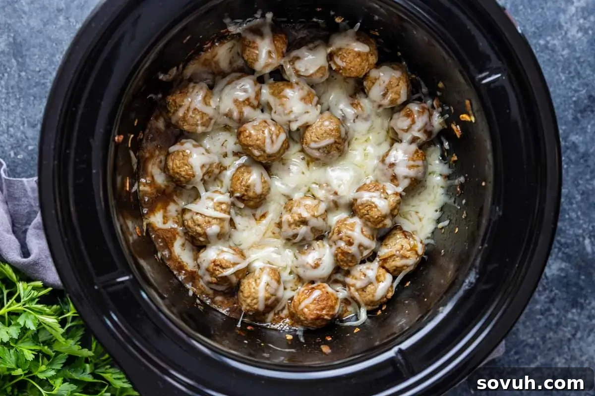 Cooked crockpot French onion meatballs topped with melted cheese inside a black slow cooker, with some parsley visible on the side—an easy appetizer meatballs recipe perfect for gatherings.