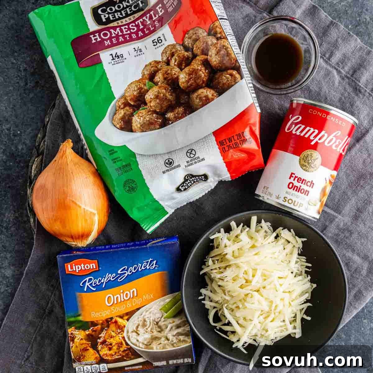 Frozen meatballs, a can of French onion soup, a yellow onion, onion soup mix, shredded cheese, and a small bowl of sauce arranged on a gray cloth—perfect for an easy appetizer meatballs dish or your next French onion meatballs recipe.