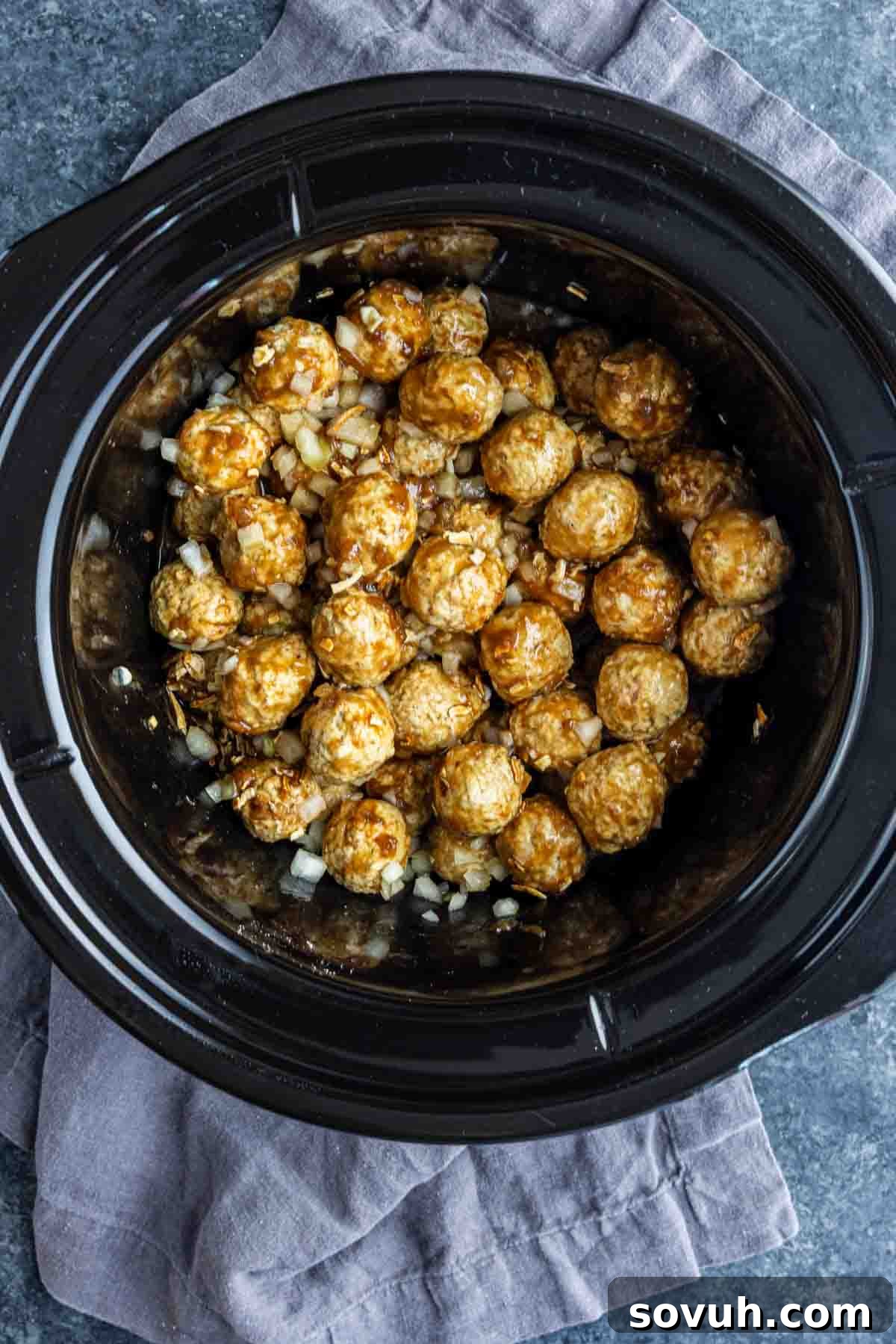 Uncooked frozen meatballs mixed with chopped onions and French onion soup mix inside a black slow cooker, placed on a gray cloth on a dark countertop.