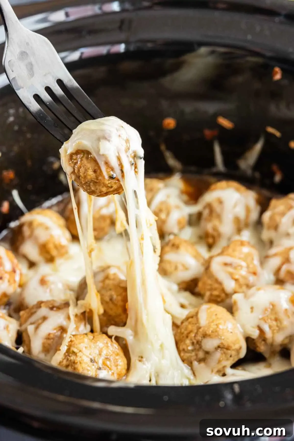 A fork lifts a cheese-covered meatball from a slow cooker filled with melted cheese and more meatballs—these crockpot French onion meatballs make an easy appetizer everyone will love.