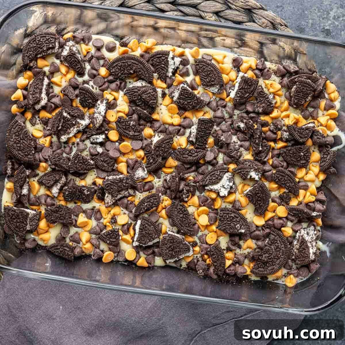 Oreo Magic Bars in a glass baking dish, adorned with chocolate chips, butterscotch chips, and Oreo chunks, poised for baking.