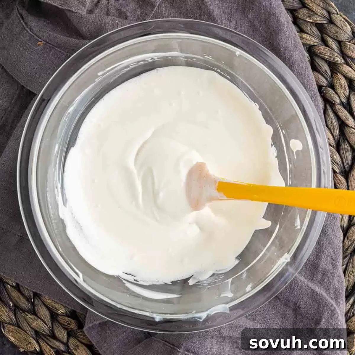 A bowl of creamy, sweetened sour cream topping for mini cheesecakes, with a yellow spoon resting inside.