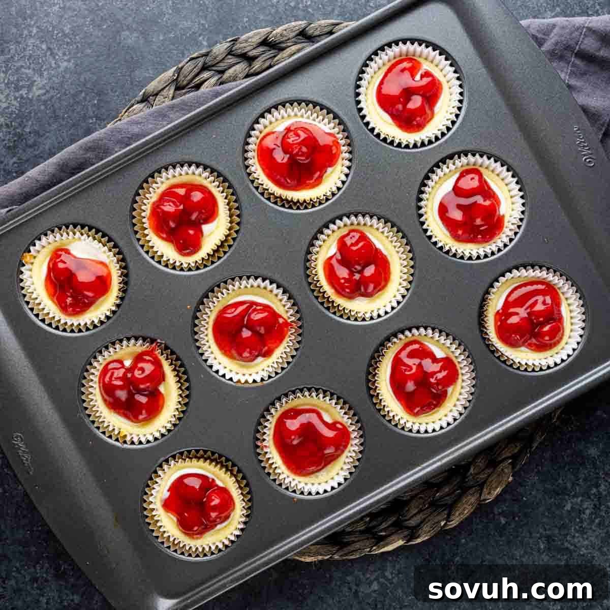 A muffin tin filled with freshly baked mini cherry cheesecakes, topped with bright red cherry pie filling.