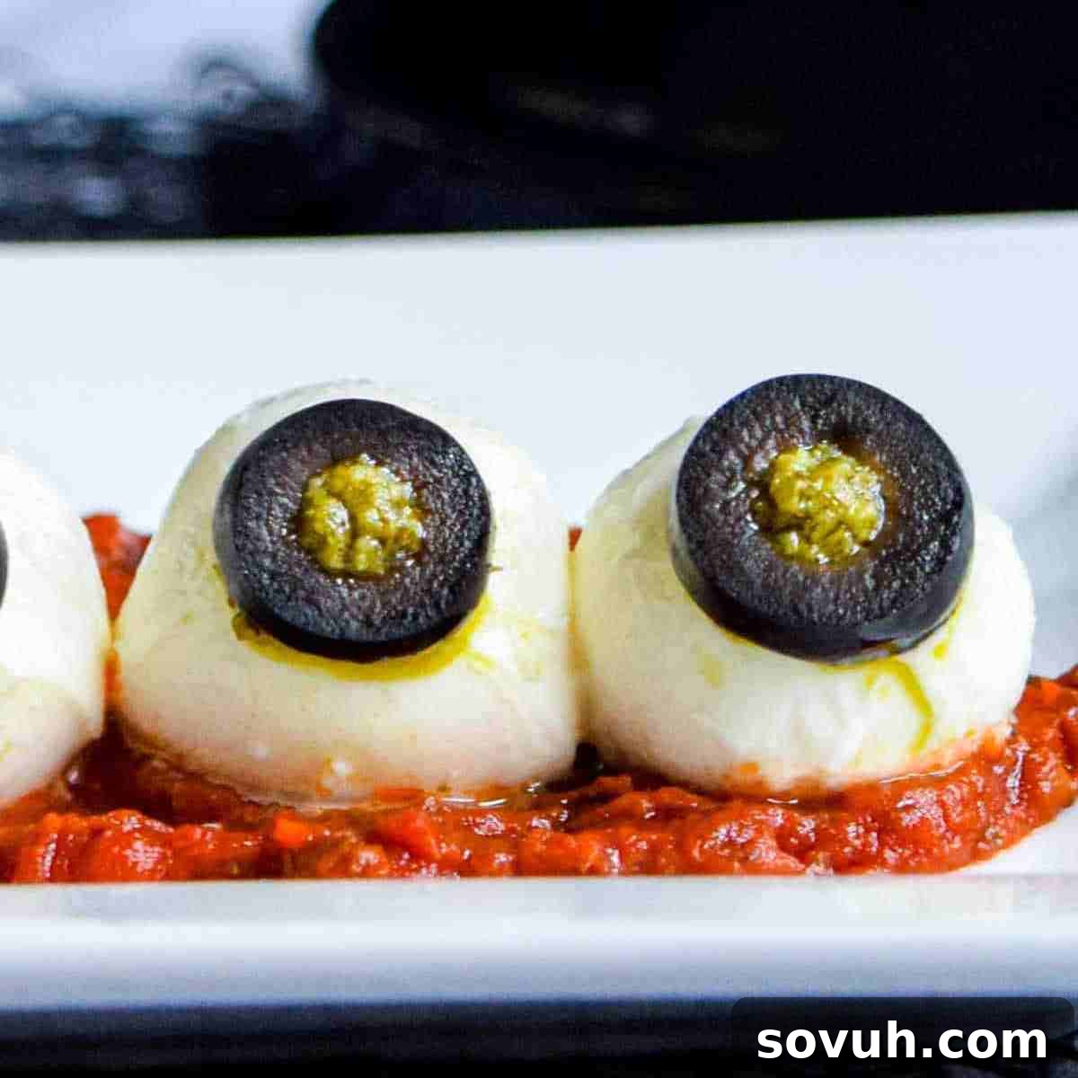 Three mozzarella balls with black olive eyes, resembling Halloween eyeballs, on a white plate, ready for a party.