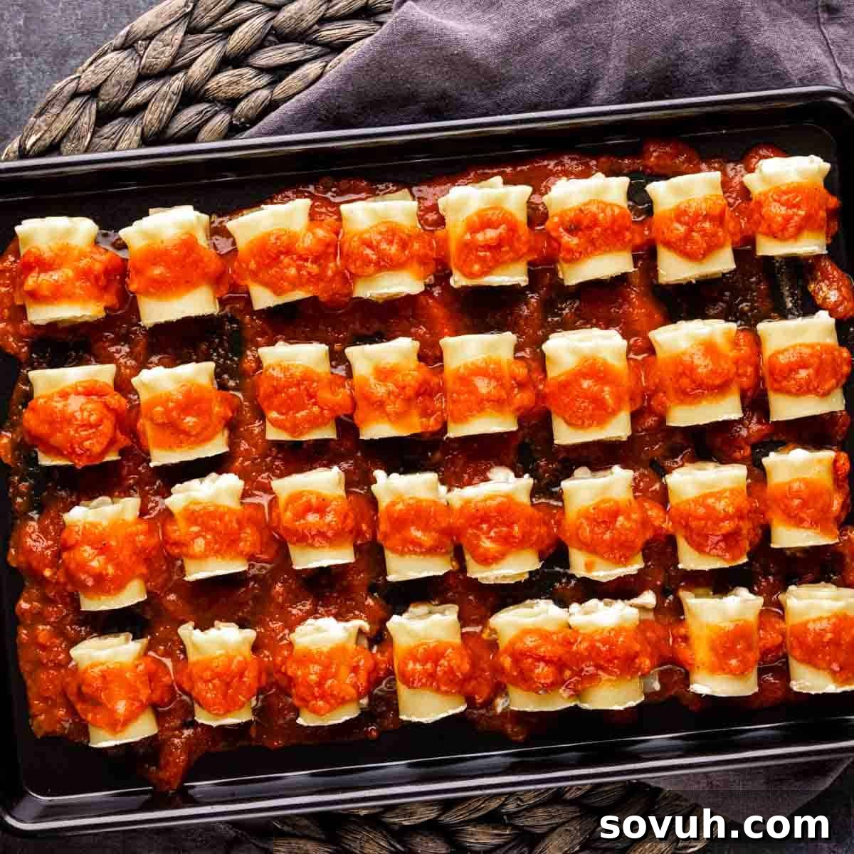 A black baking sheet lined with a thin layer of pasta sauce, ready for the arrangement of mini lasagna rolls.