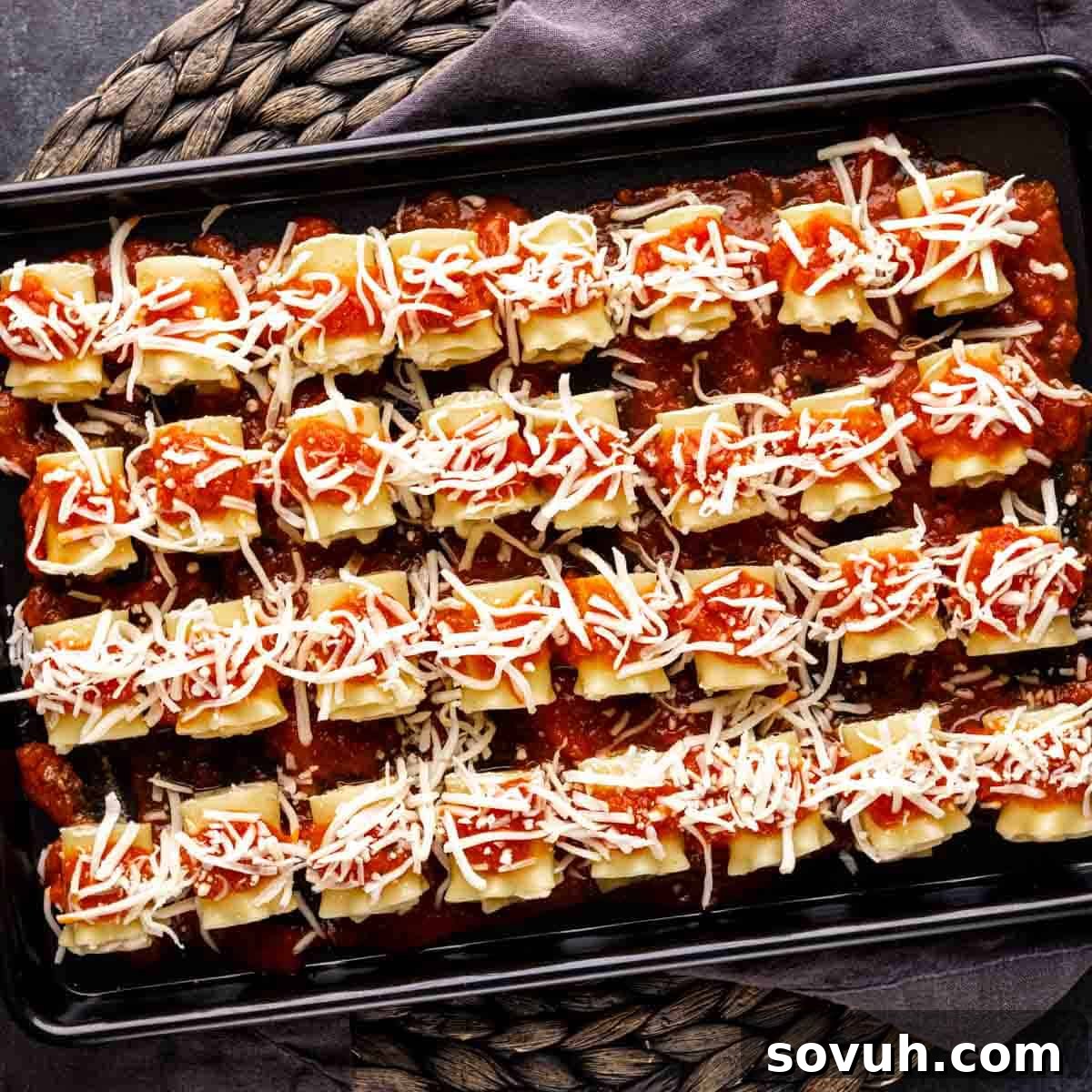 Mini lasagna rolls meticulously arranged on a baking sheet, each topped with a dollop of sauce and a generous sprinkle of shredded mozzarella cheese.