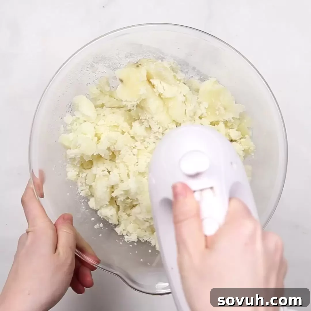 A person using a hand mixer to mash potatoes in a bowl for Duchess Potatoes, aiming for a perfectly smooth texture.