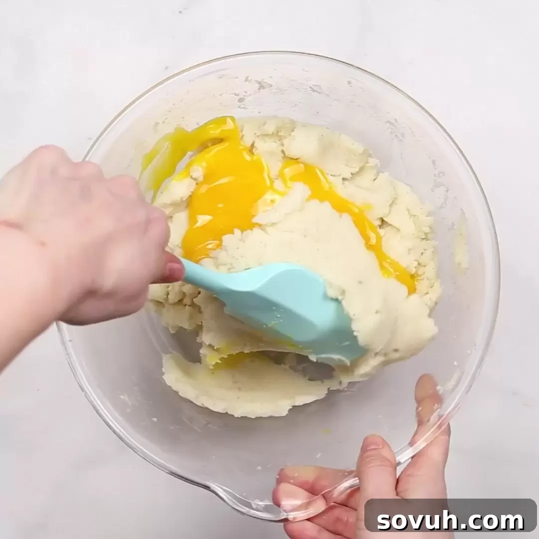 A person mixing ingredients in a bowl with a blue spatula for Duchess Potatoes, gently folding in egg yolks.