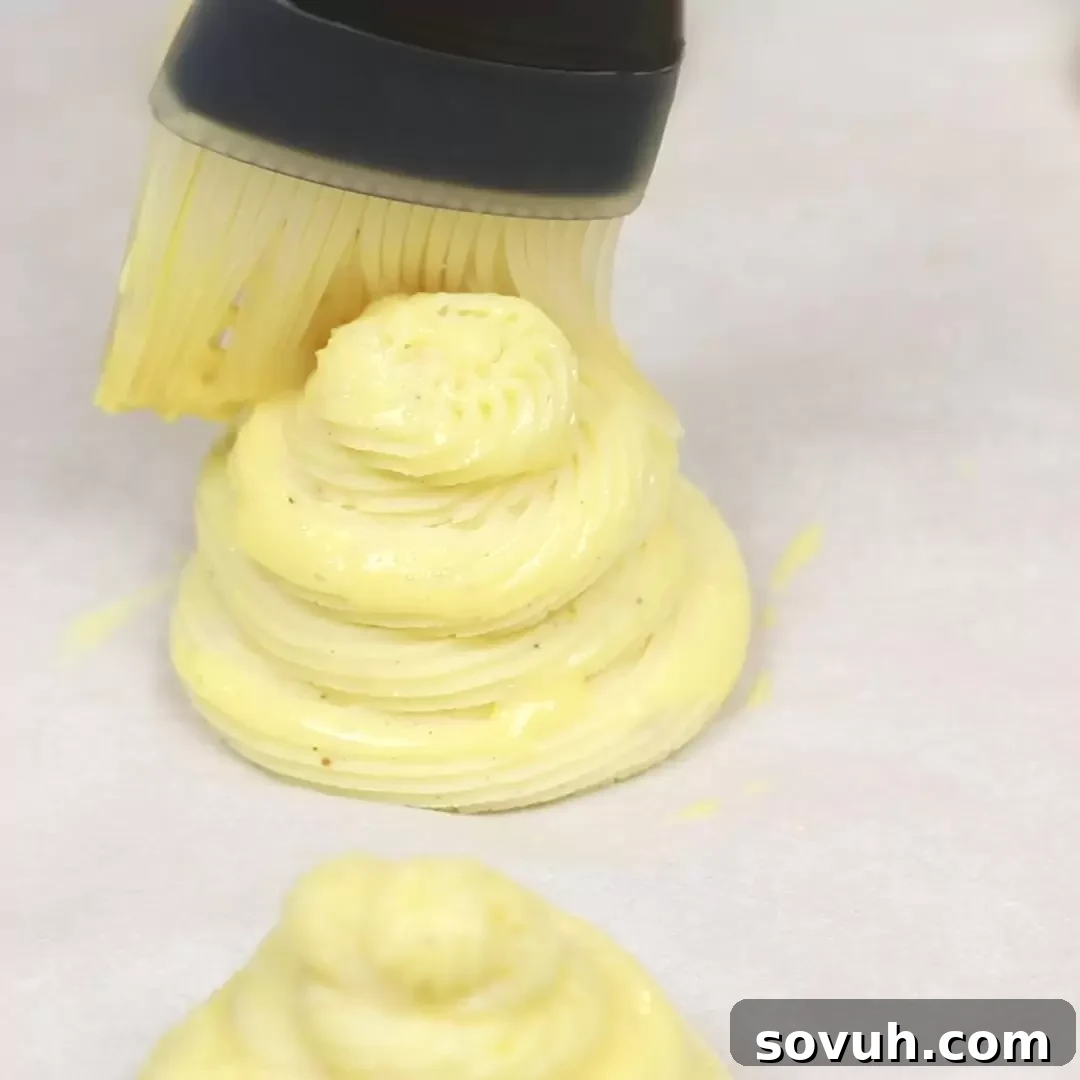 A person is using a brush to apply egg wash on Duchess Potatoes, ensuring a golden-brown finish.
