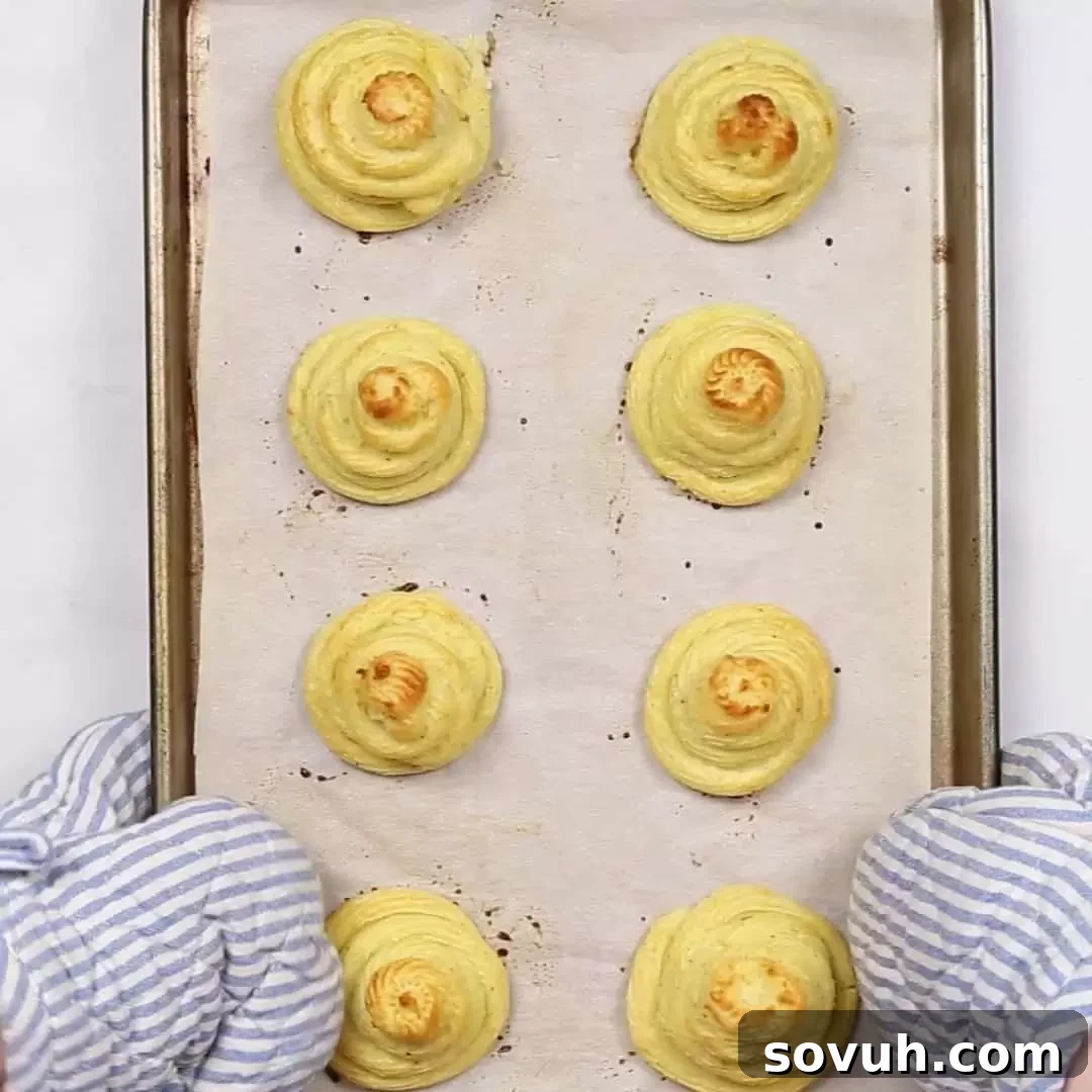 A person holding a baking sheet full of golden-brown baked Duchess Potatoes, fresh from the oven.