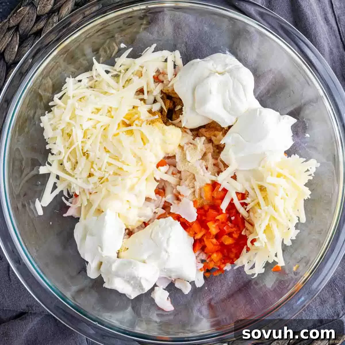 A glass bowl filled with a creamy mixture of cheese, finely chopped red bell pepper, and various spices, ready for the crab meat.