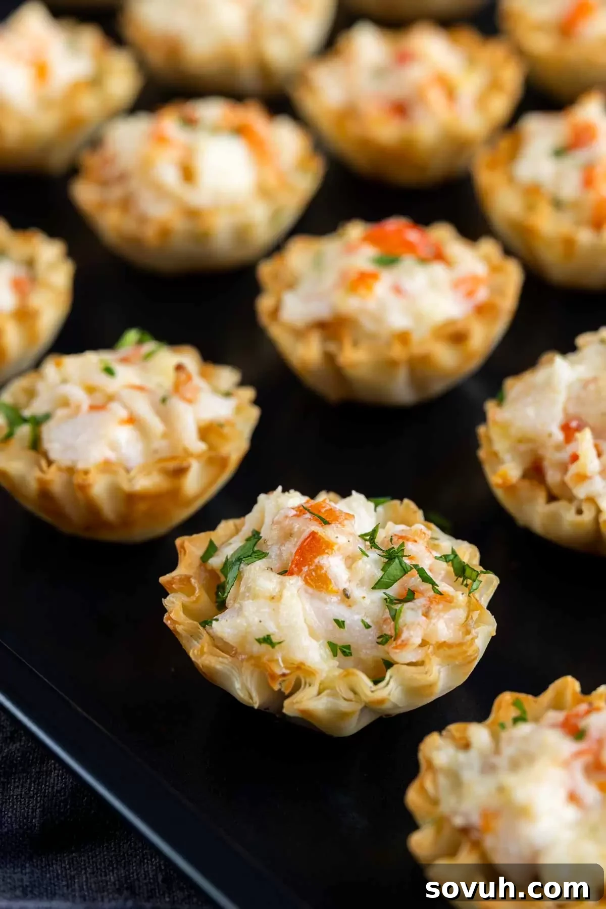 A close-up of a tray of mini crab puffs, showing the creamy filling, melted cheese, and fresh parsley garnish.