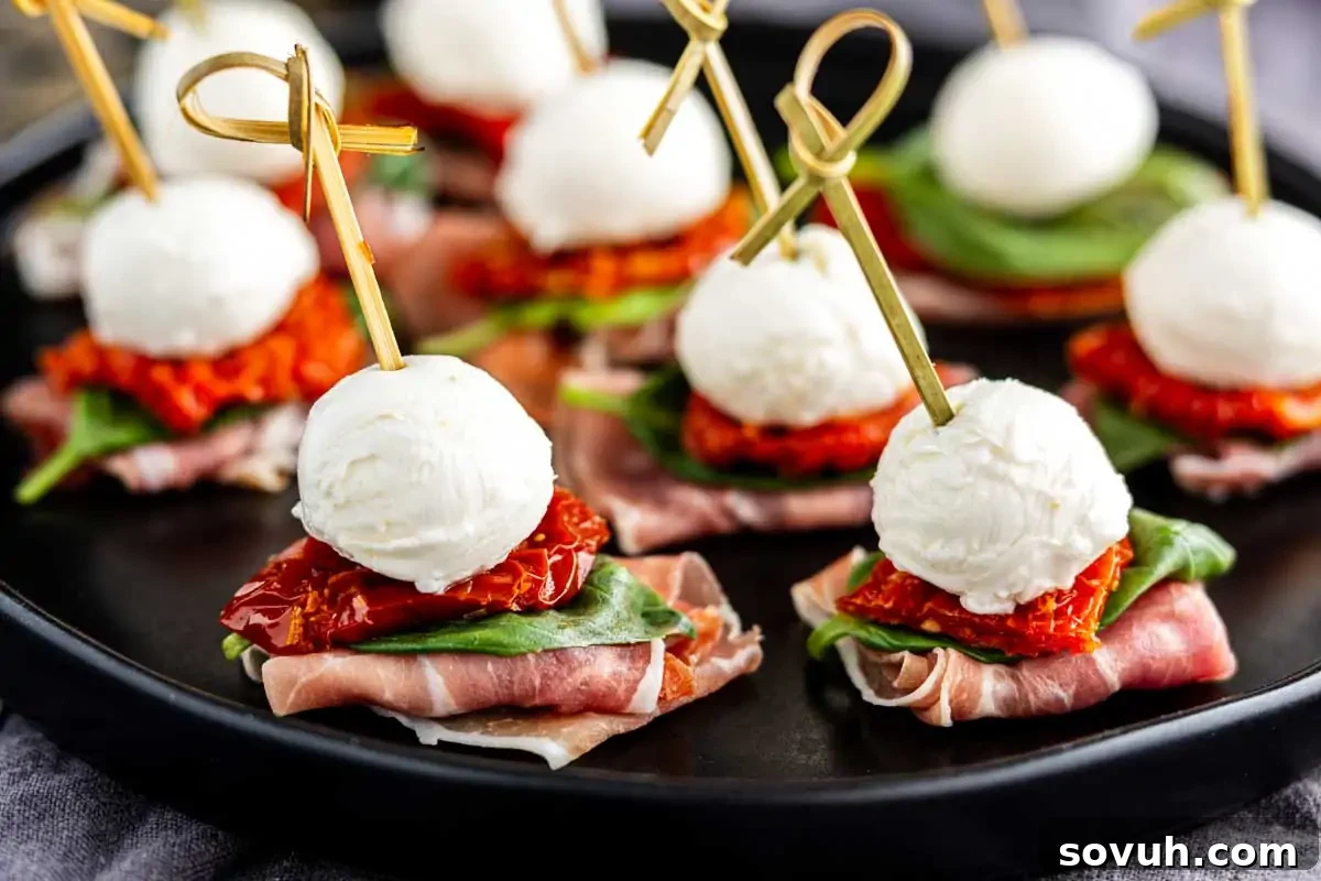 Appetizers on skewers with fresh mozzarella balls, vibrant sun-dried tomatoes, tender spinach, and savory prosciutto, elegantly arranged on a sleek black plate.