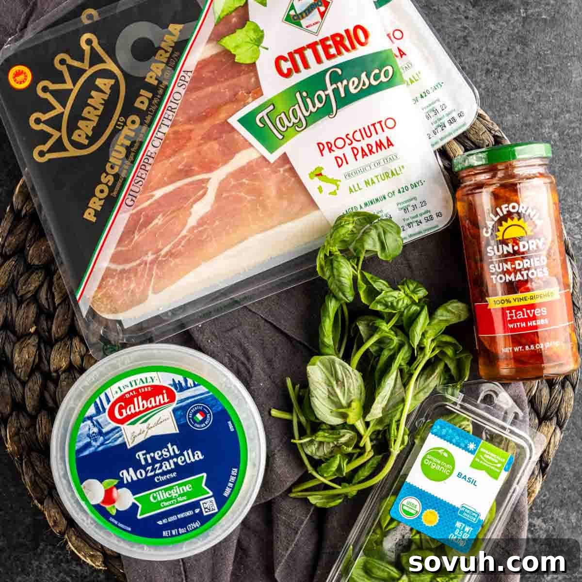 An assortment of premium Italian ingredients including thinly sliced prosciutto, fresh ciliegine mozzarella, vibrant sun-dried tomatoes, and fragrant basil leaves on a dark textured surface, ready for assembly.