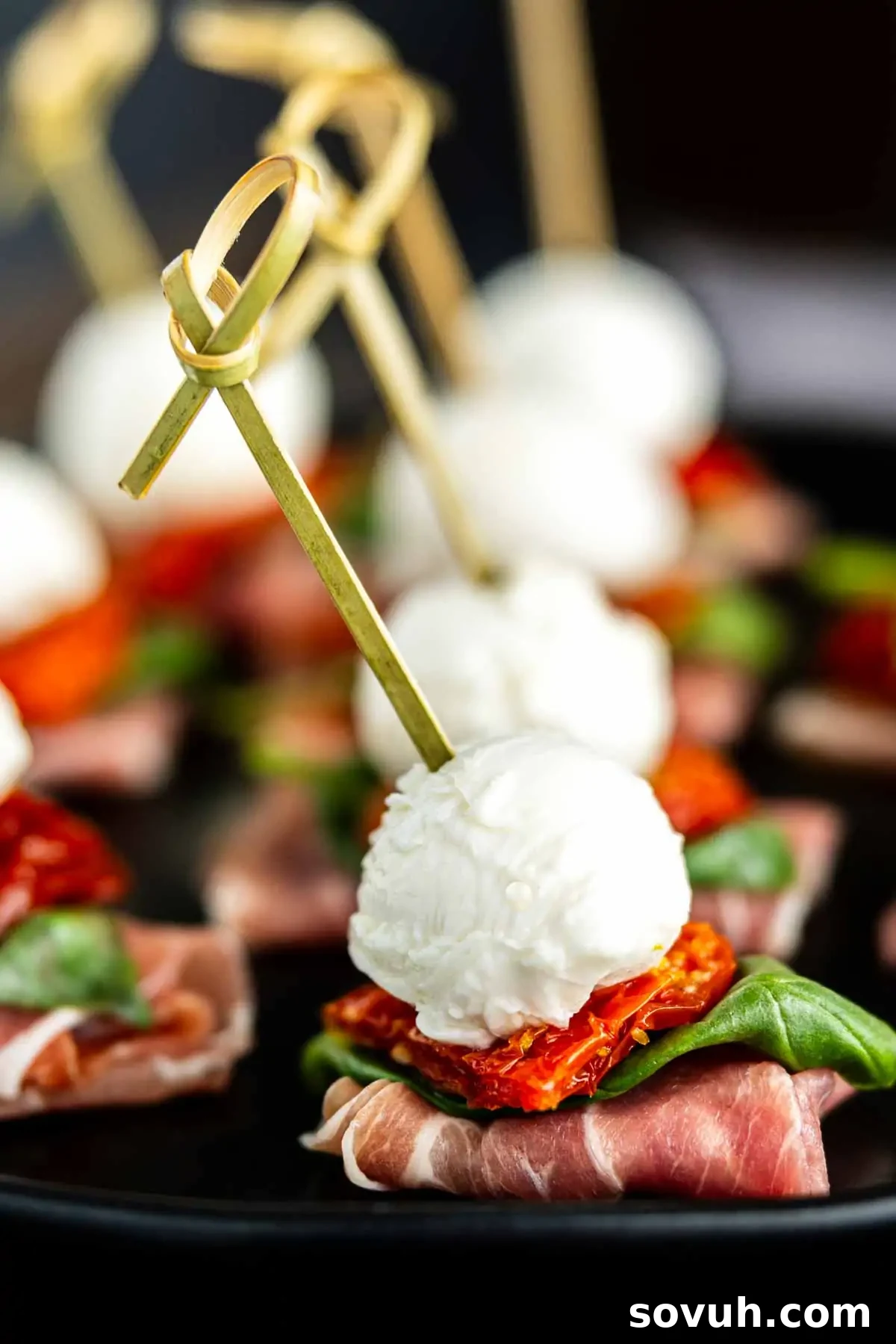 Exquisite Caprese-style antipasto skewers featuring creamy mozzarella balls, vibrant sun-dried tomatoes, and fresh basil leaves, all wrapped in delicate prosciutto slices and artfully arranged on a dark serving tray.