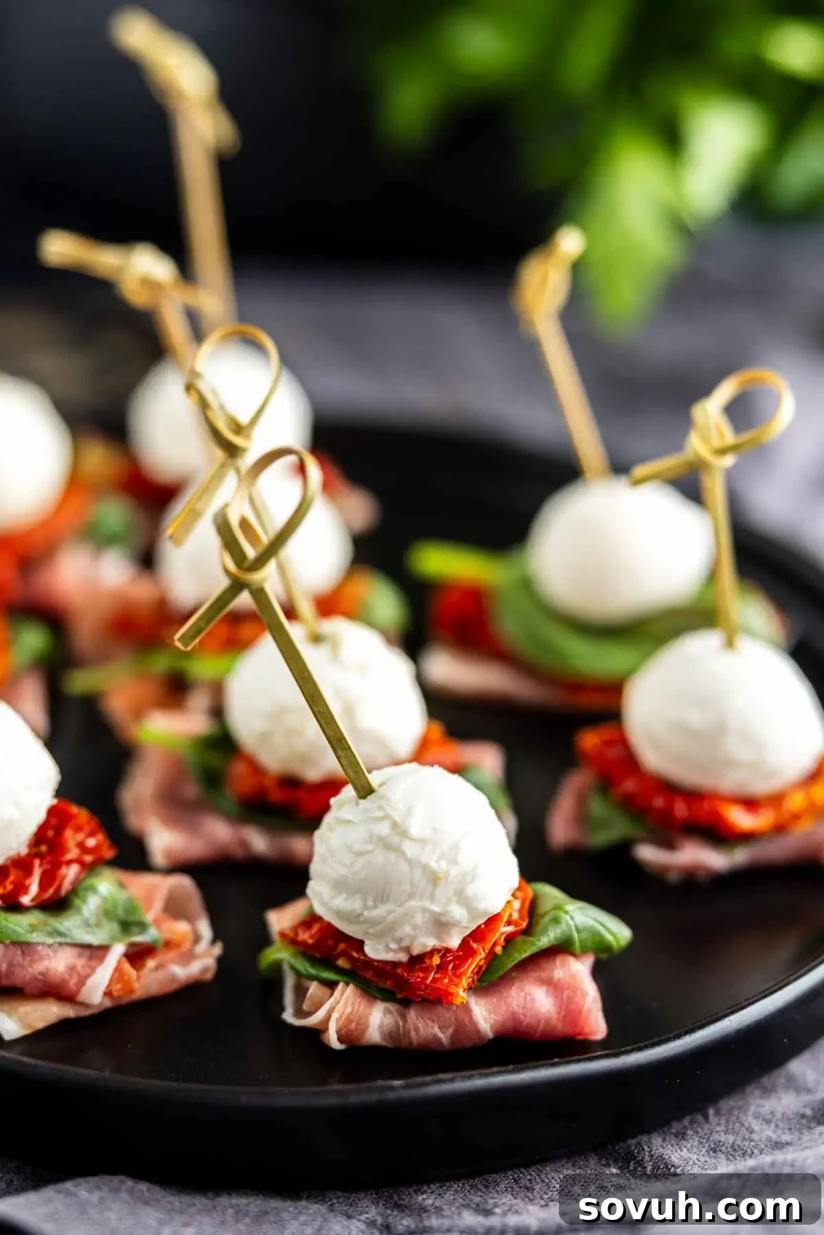 Appetizers on a black plate featuring skewers with mozzarella balls, fresh basil, sweet sun-dried tomatoes, and savory prosciutto, artfully arranged for serving.