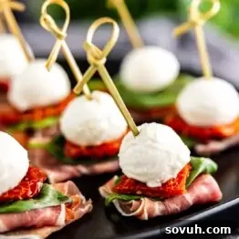 Caprese skewers with mozzarella, basil, and tomato on prosciutto slices, presented on a black plate.