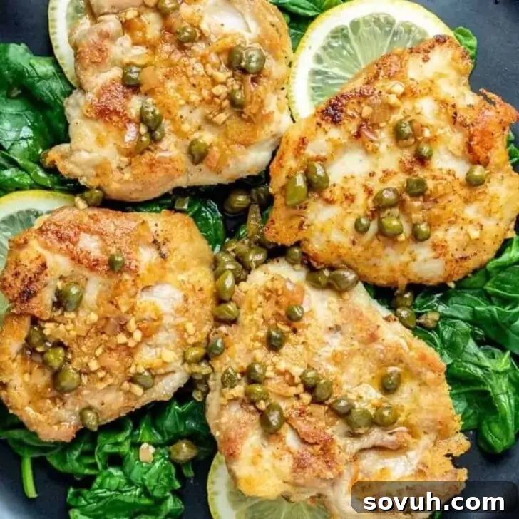 Four pieces of pan-seared chicken topped with capers and garlic, served on a bed of sautéed spinach and garnished with fresh lemon slices.