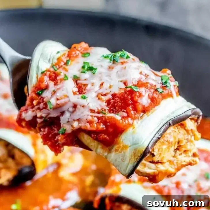 A close-up of a fork holding a piece of cheesy, sauce-covered eggplant rollatini, highlighting the shredded chicken filling. The pan filled with more rollatini is in the background, garnished with chopped herbs.