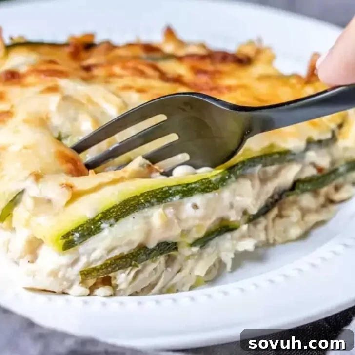 A piece of layered casserole with cheese and zucchini being cut with a black fork on a white plate, revealing creamy layers.