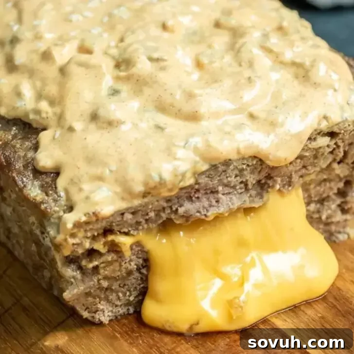 A close-up view of a Big Mac Keto Meatloaf topped with a creamy sauce, sliced to reveal layers of melted cheese oozing out onto a wooden cutting board, garnished with pickles.