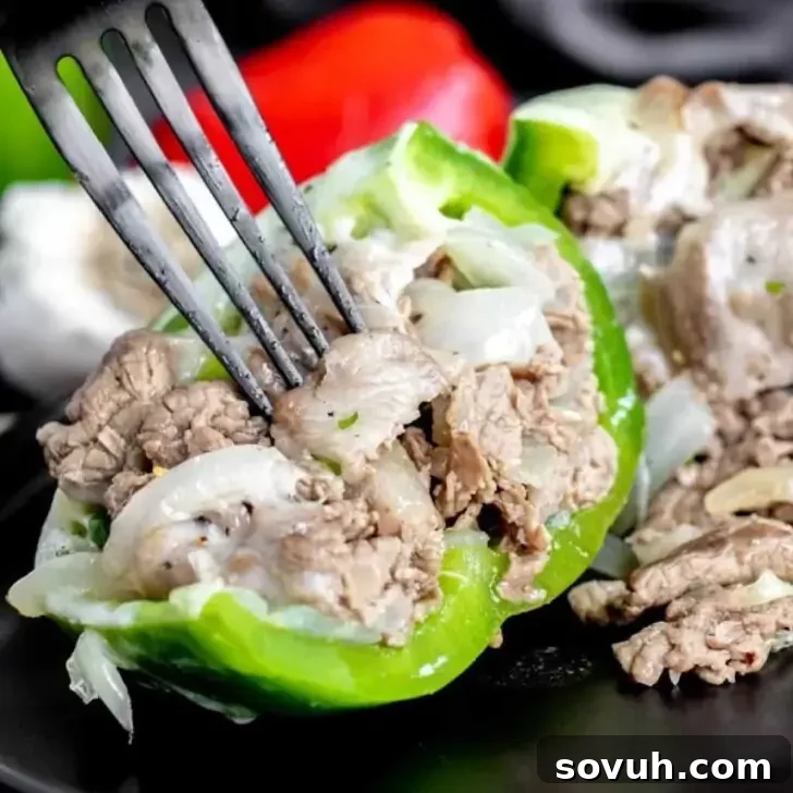 Close-up of a fork holding a piece of cooked beef and onion mixture inside a halved green bell pepper, with more Philly Cheesesteak Stuffed Peppers in the background.