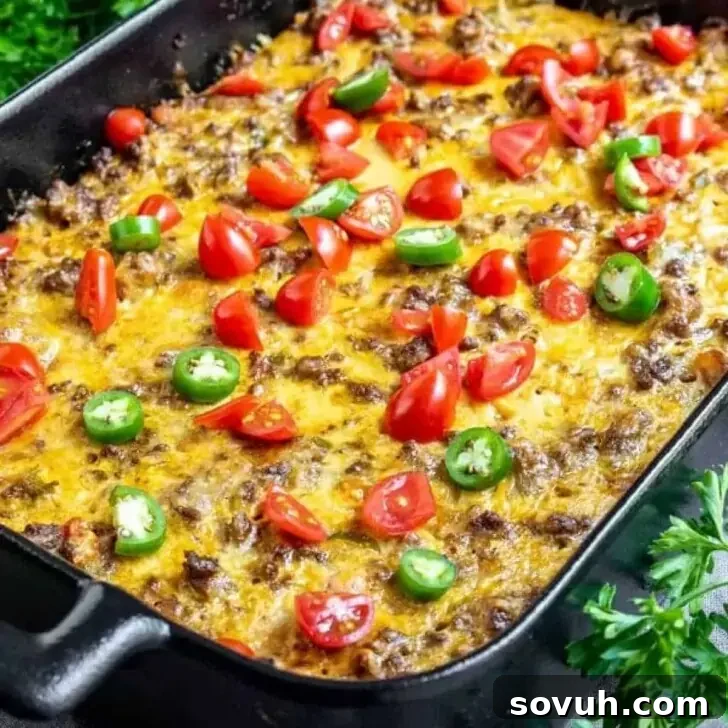 A baked casserole topped with melted cheese, browned ground meat, sliced fresh tomatoes, and green jalapeño pieces in a black baking dish, garnished with fresh parsley.