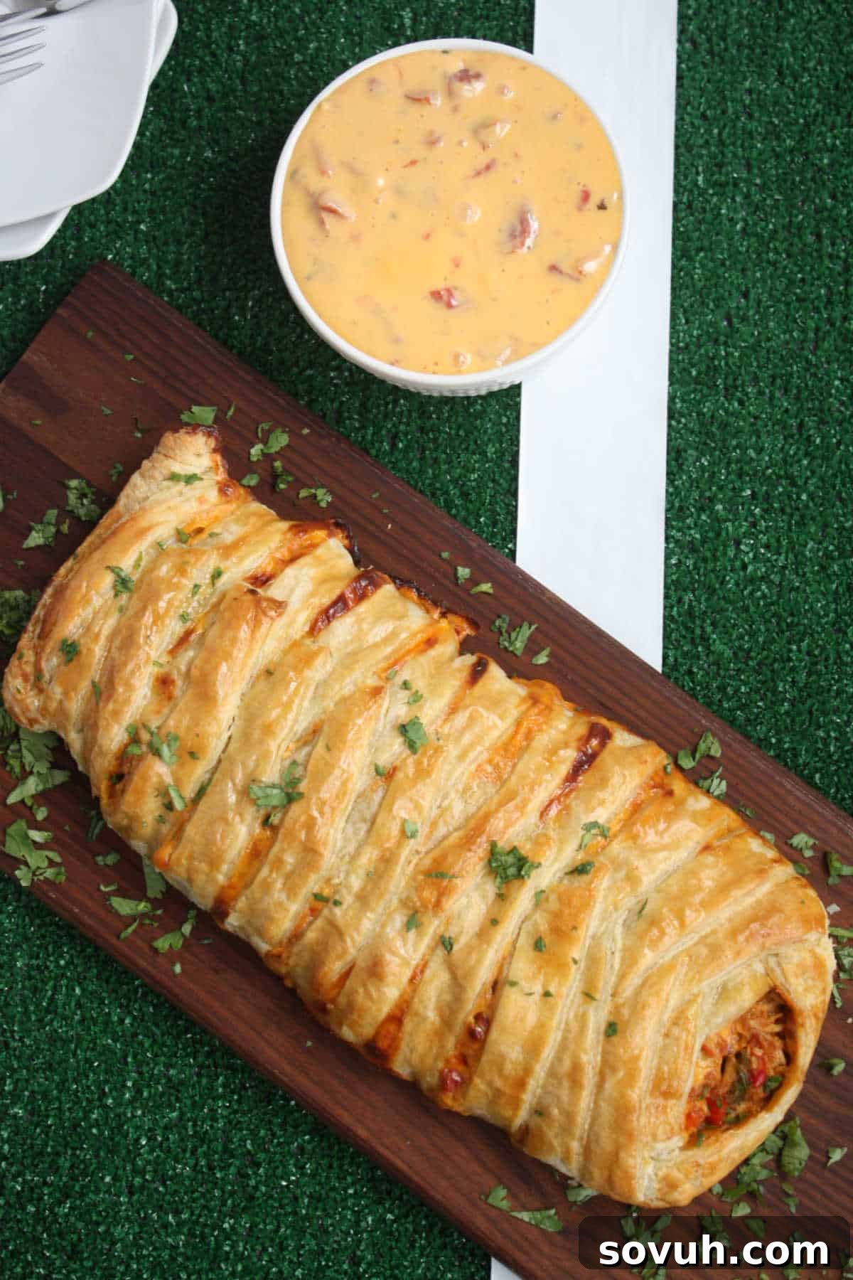 A braided puff pastry filled with a cooked mixture sits on a wooden board, garnished with chopped herbs. A bowl of cheese dip with visible chunks is placed nearby on a green, field-like surface.
