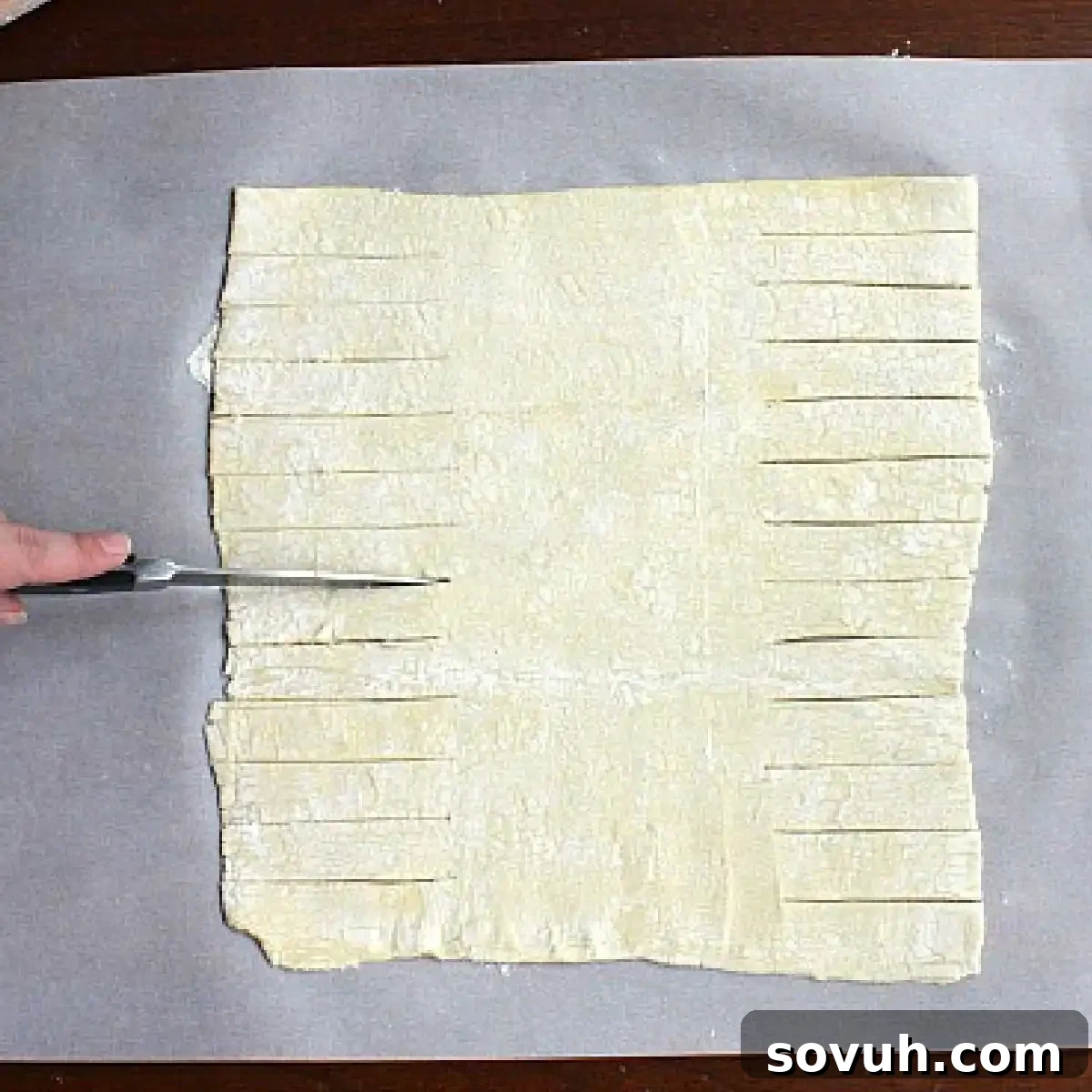 A hand precisely cutting strips into a sheet of thawed puff pastry dough laid out on parchment paper, preparing it for the taco filling.