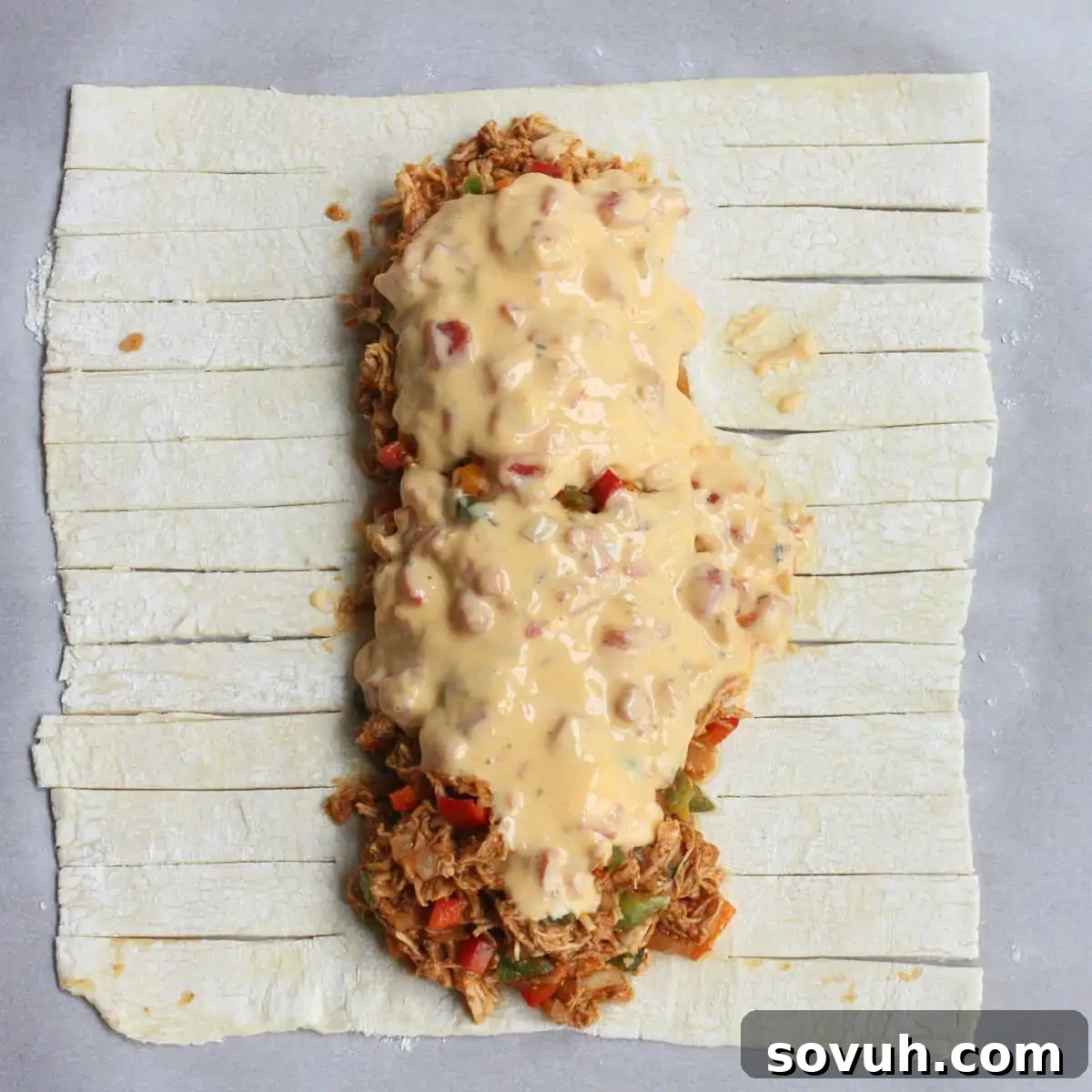 An uncooked puff pastry sheet, neatly divided into strips, with a generous line of savory shredded chicken mixture and creamy cheese sauce placed down the center.