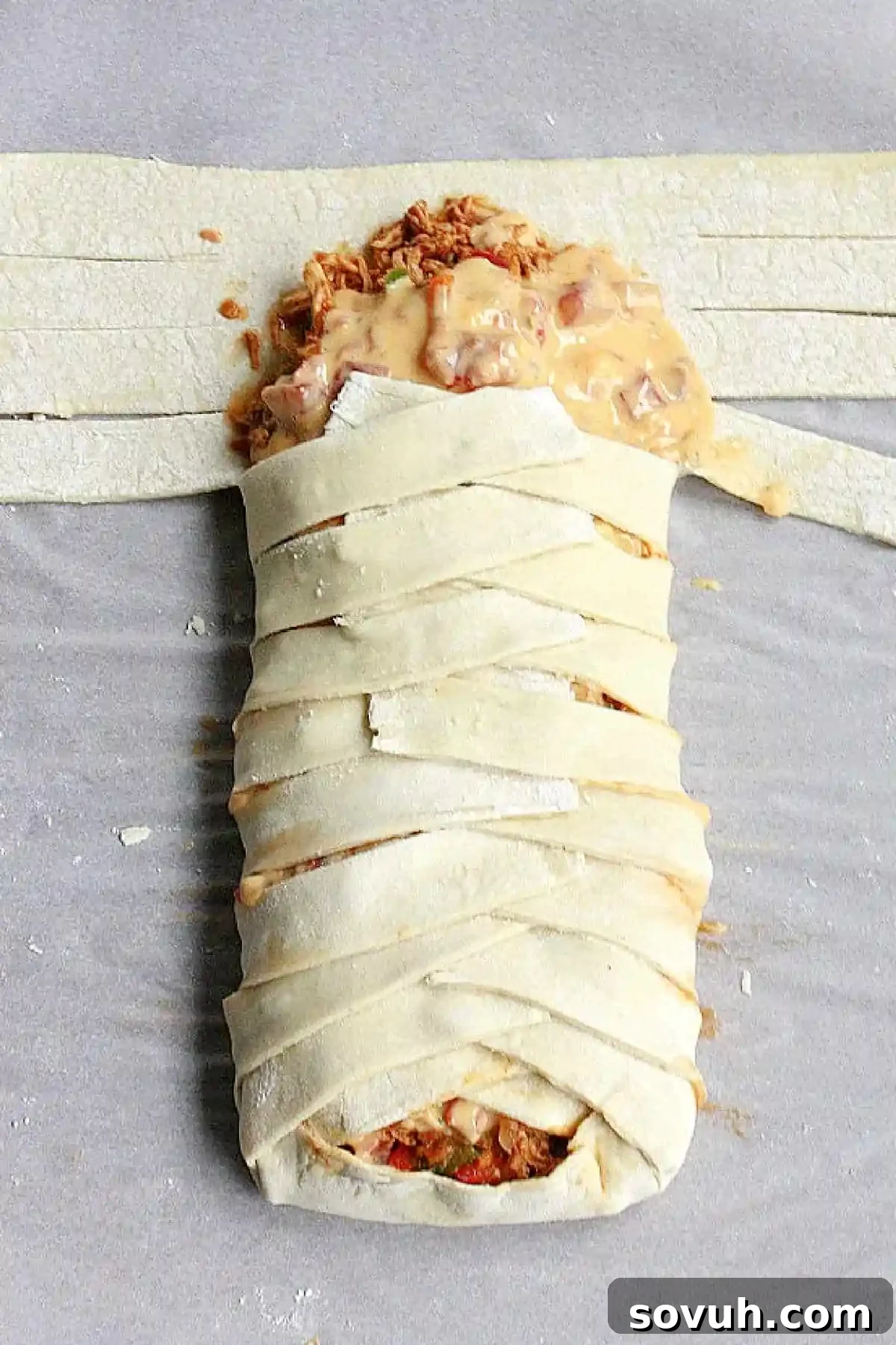 A partially braided puff pastry, filled with chicken and cheese, with some strips folded over and others still waiting, on a parchment-lined baking sheet.