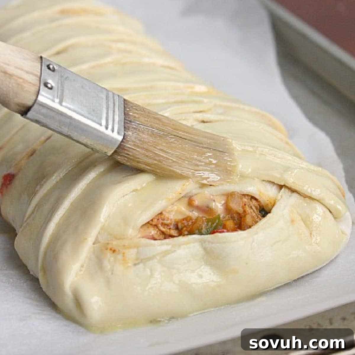 A pastry brush applying a glossy egg wash to an uncooked chicken taco braid, enhancing its future golden crust.