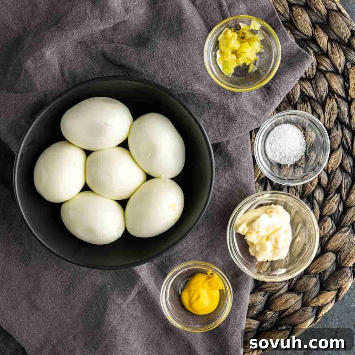 The Devil's Own Pickles 3 A dark ceramic bowl holds six perfectly hard-boiled eggs, ready for preparation. Surrounding it are small, clear glass bowls containing the key ingredients for making delightful deviled eggs with a pickle twist, or easy pickle appetizers: finely chopped pickles, a pinch of sea salt, creamy mayonnaise, and bright yellow mustard. All are arranged neatly on a grey linen cloth, accented by a woven placemat, highlighting the fresh ingredients.