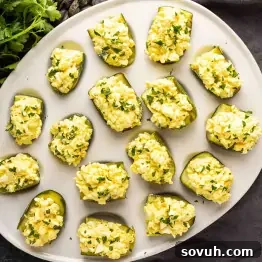 A white plate featuring pickle halves generously topped with a creamy egg salad mixture, elegantly garnished with chopped fresh herbs.