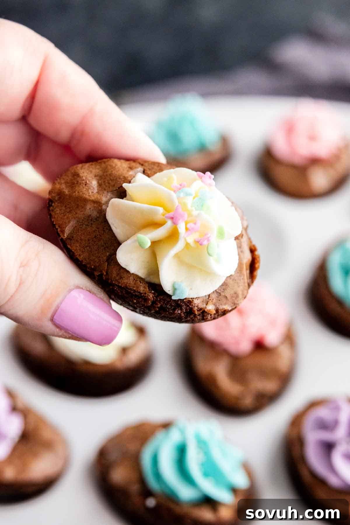 Devilish Easter Brownies from a Box 6 A hand with painted nails holds a small chocolate cookie topped with white frosting and pastel star sprinkles; more easy Easter dessert cookies made from brownie mix are on a plate in the background.