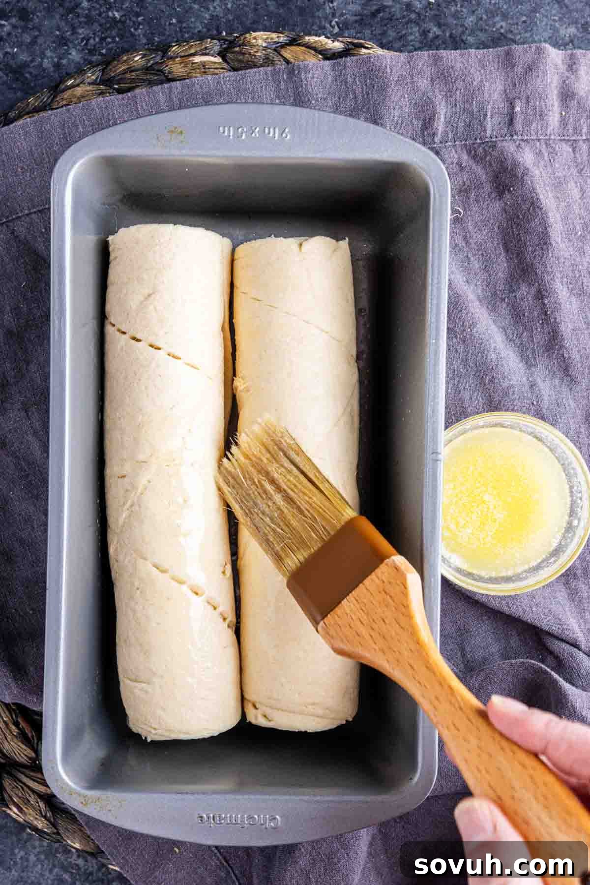 The Viral Golden Crescent Bake 4 A hand uses a pastry brush to apply melted butter to two unbaked Viral TikTok Crescent Roll Loaf dough rolls in a loaf pan on a dark surface, preparing for baking.