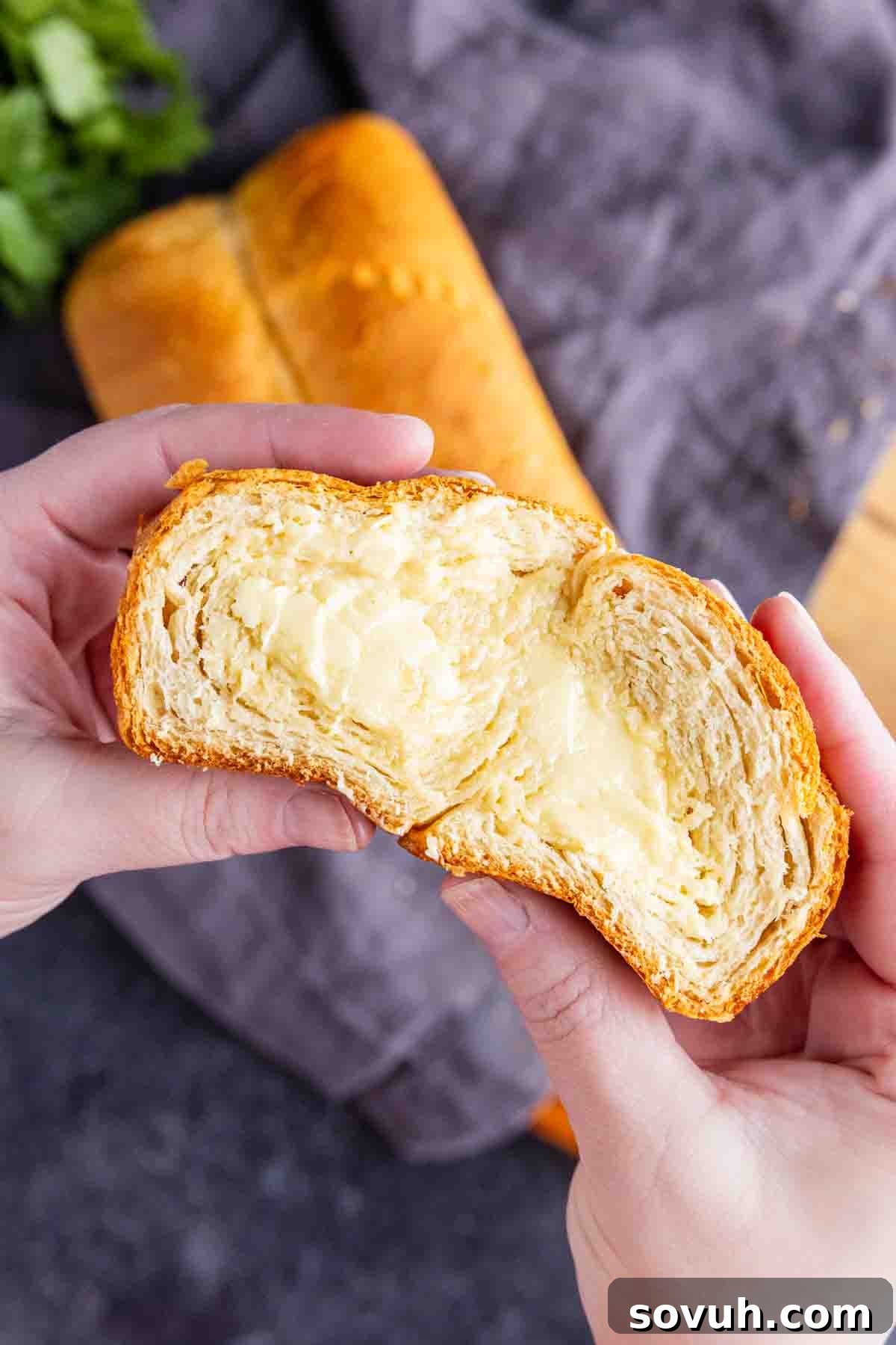 The Viral Golden Crescent Bake 7 Two hands hold a crescent roll bread split in half, revealing its flaky layers and creamy filling, with a buttery bread loaf and a gray cloth in the background, showcasing its delicious texture.