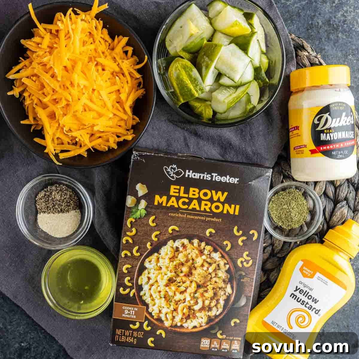 Quick and Zesty Dill Pickle Pasta Salad 3 A vibrant flat lay of ingredients for a Dill Pickle Pasta Salad on a rustic wooden surface: golden elbow macaroni, a mound of shredded sharp cheddar cheese, bright green chopped dill pickles, creamy mayonnaise, zesty yellow mustard, a bottle of clear oil, black pepper, and fragrant dried dill. These simple yet essential components are ready to transform into a delicious and easy side dish.