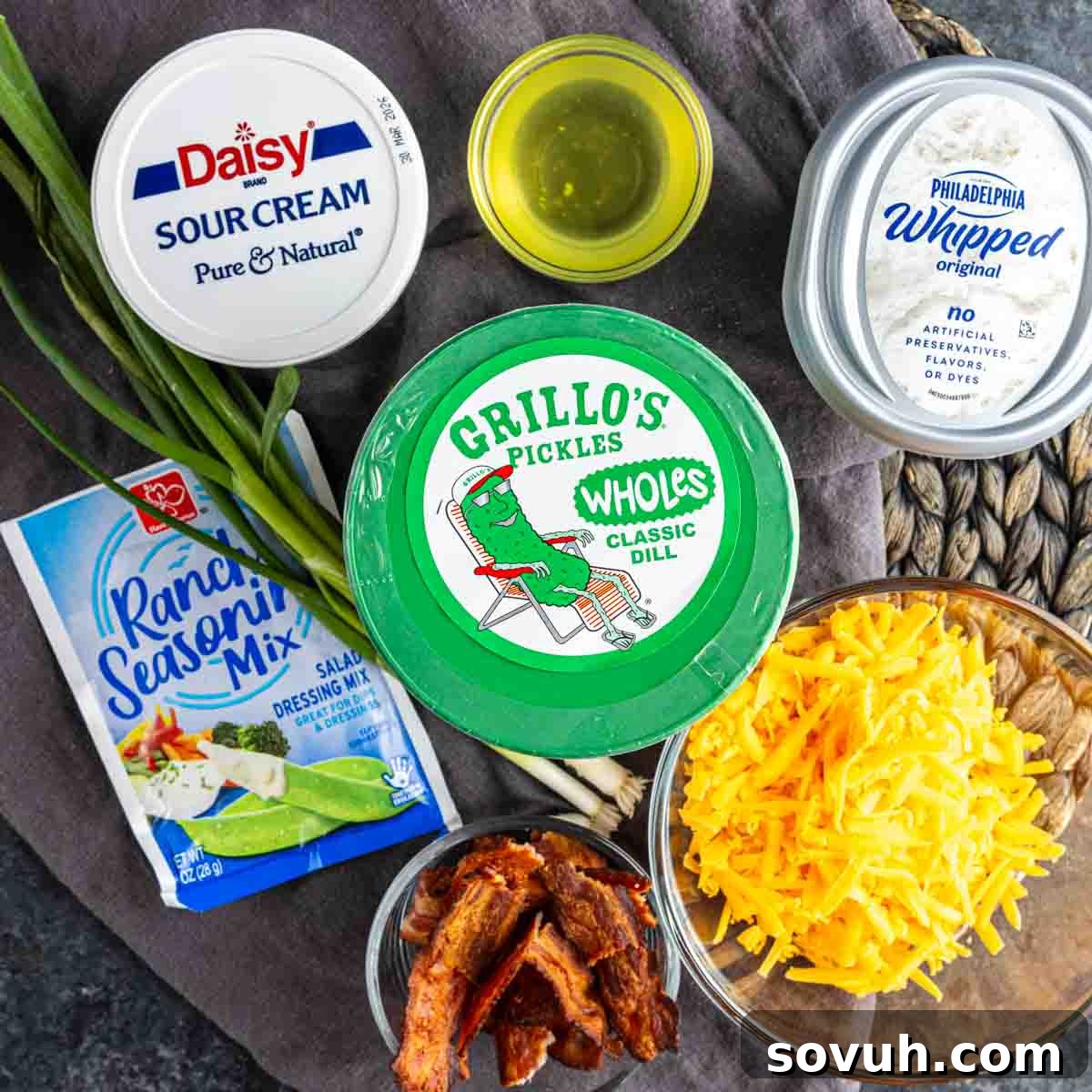 Your New Obsession: The Viral Pickle Dip Recipe 3 Assorted ingredients displayed on a table, ready to make the viral pickle dip: sour cream, whipped cream cheese, ranch seasoning mix, cheddar cheese, cooked bacon, green onions, pickles, and a small bowl of oil.