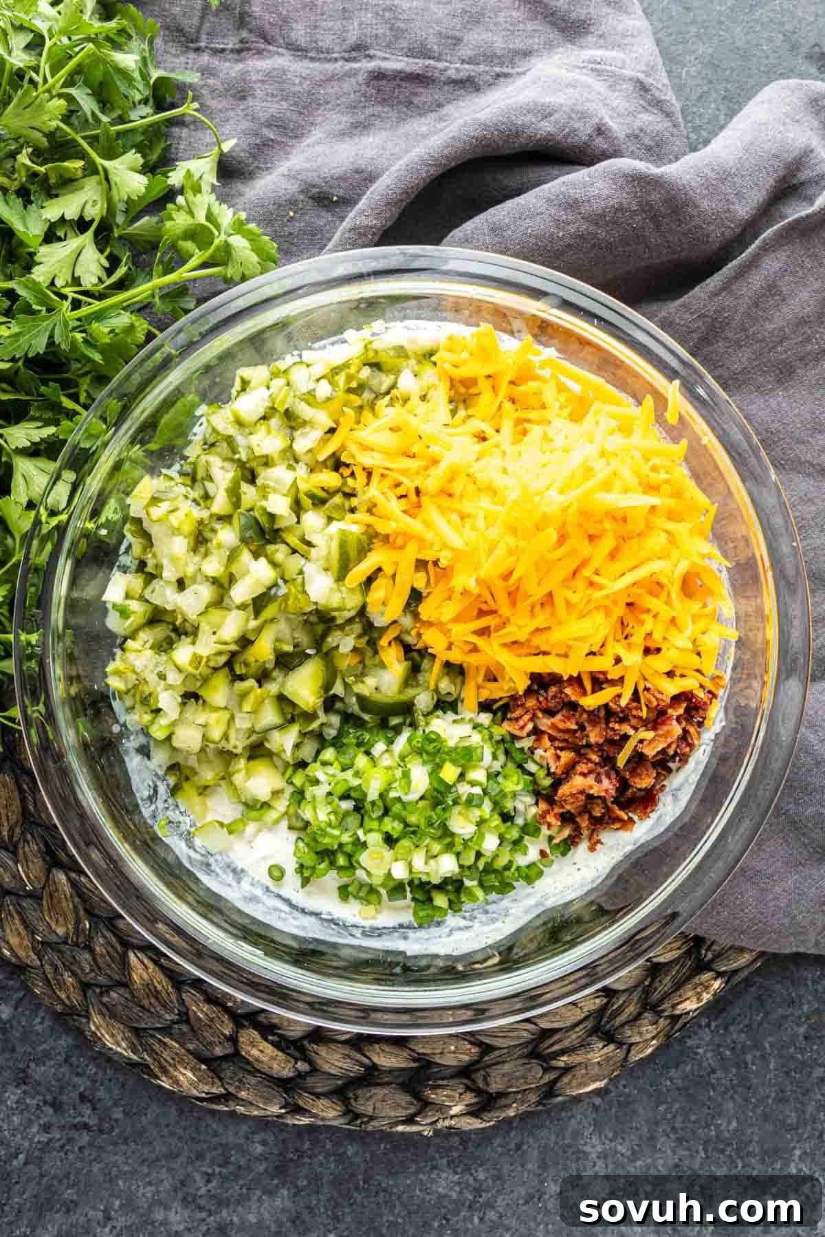 Your New Obsession: The Viral Pickle Dip Recipe 4 A glass bowl with chopped pickles, shredded cheddar cheese, green onions, and bacon bits on a creamy base—this cold party dip, inspired by the Viral Pickle Dip trend on TikTok, sits next to fresh cilantro on a dark surface.