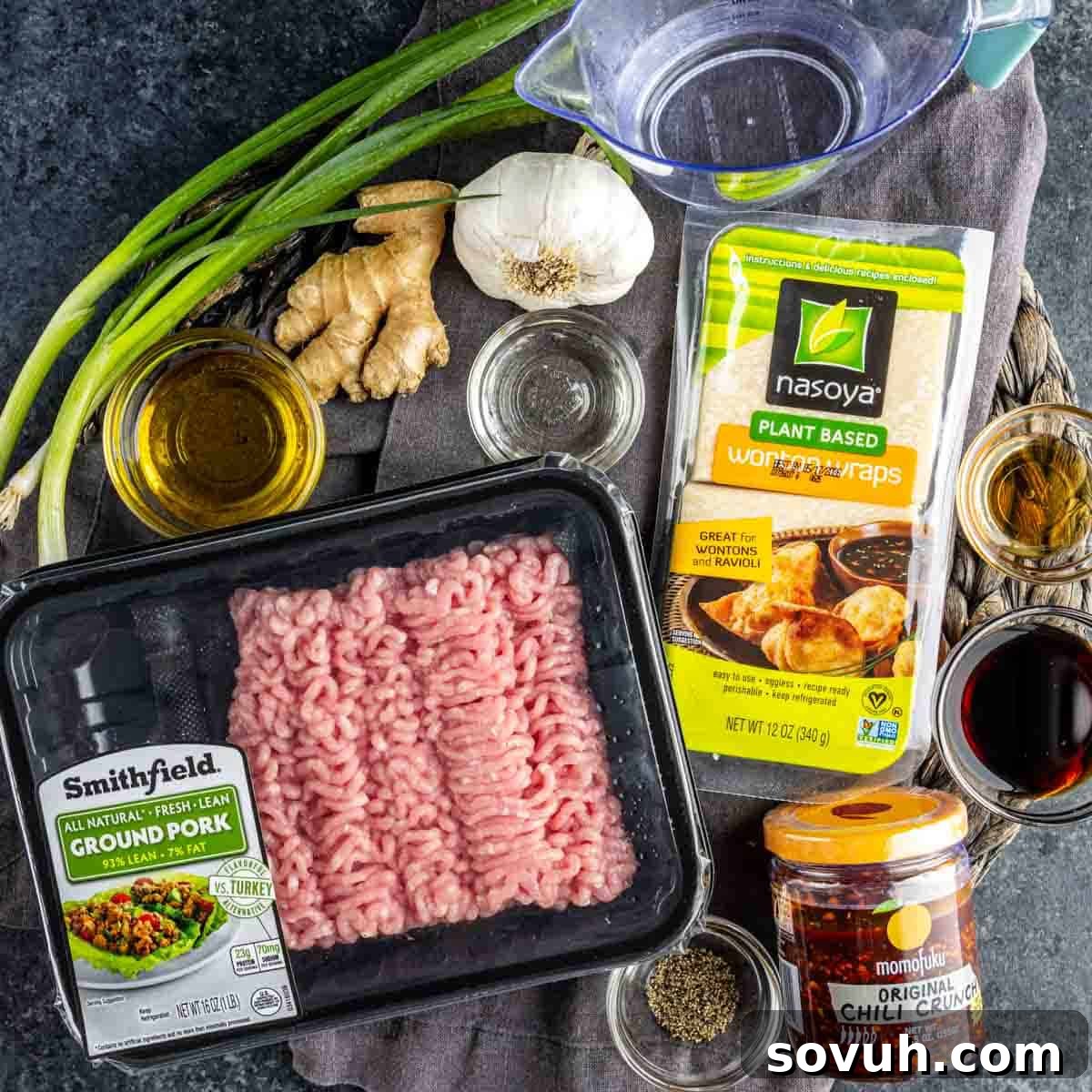 Effortless Viral Blanket Dumplings No Folding Needed 3 Various ingredients are arranged on a table, including ground pork, wonton wraps for TikTok dumplings, chili crunch, green onions, ginger, garlic, seasonings, and a measuring cup with water.